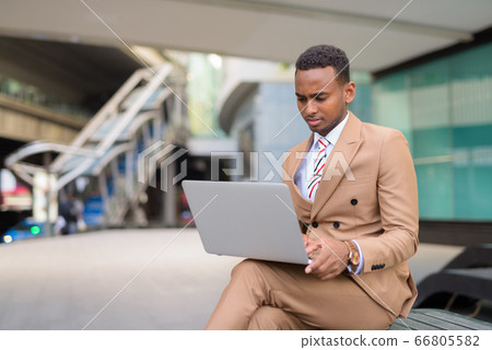 Young handsome African businessman using laptop while sitting in the city outdoors Young handsome African businessman using laptop while sitting in the city outdoors 66805582