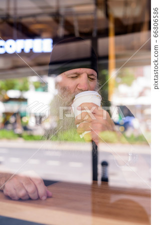 Mature handsome bearded hipster man drinking coffee at the coffee shop through the window Mature handsome bearded hipster man drinking coffee at the coffee shop through the window 66806586