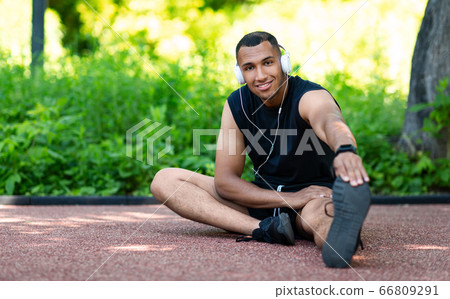 Portrait of young black man in headphones stretching his legs on jogging track at park, copy space 66809291