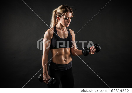 Fitness in gym, sport and healthy lifestyle concept. Beautiful athletic woman showing her trained body on black background. Bodybuilder female model training biceps muscles with dumbbell. 66819260