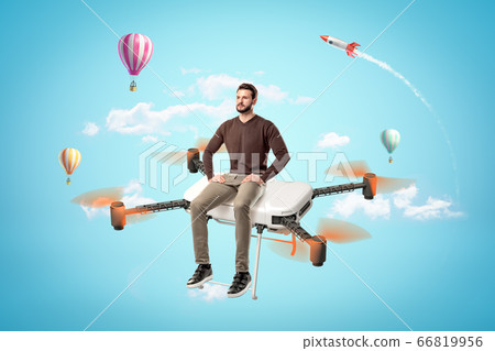 Serious handsome young man in casual clothes sitting on big quadcopter in blue sky with hot air balloons and space rocket in background. Serious handsome young man in casual clothes sitting on big quadcopter in blue sky with hot air balloons and space rocket in background. 66819956