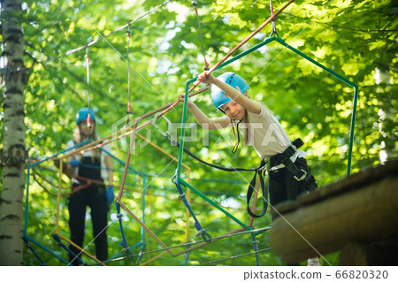 Rope adventure - a little girl walks on the rope bridge and holding by the ropes with her hands - her mother watching her Rope adventure - a little girl walks on the rope bridge and holding by the ropes with her hands - her mother watching her 66820320