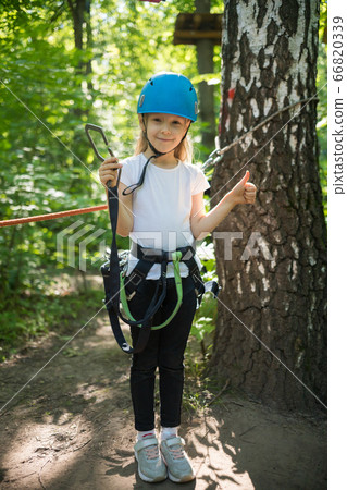Rope adventure - a little girl with an insurance belt standing in the forest and holding an insurance hook Rope adventure - a little girl with an insurance belt standing in the forest and holding an insurance hook 66820339