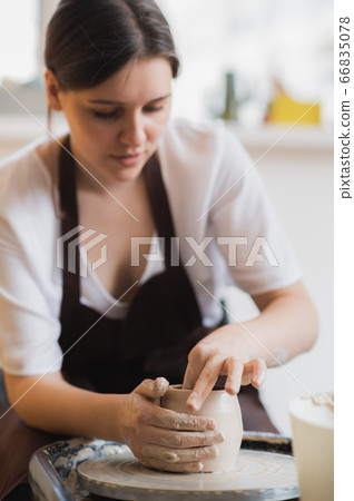 Closeup of young female potter with sponge keeping it close to rotating clay pot while shaping form of pot during work 66835078