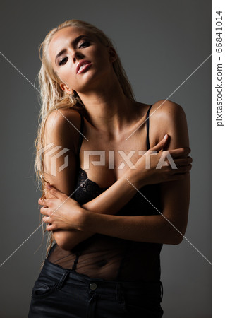 Portrait of blond young woman with beautiful makeup and hairstyle in a black lace bodysuit stands on gray background. Fashion model poses in elegant clothes in studio 66841014