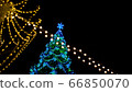 View of Christmas tree decorated large luminous multi-pointed blue stars and yellow balls through garland of yellow lights encircling city square 66850070