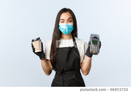 Coffee shop, coronavirus, social distancing and contactless payment concept. Cheeky cute asian waitress, barista in medical mask and gloves, wink as handing coffee order and paying terminal Coffee shop, coronavirus, social distancing and contactless payment concept. Cheeky cute asian waitress, barista in medical mask and gloves, wink as handing coffee order and paying terminal 66853720