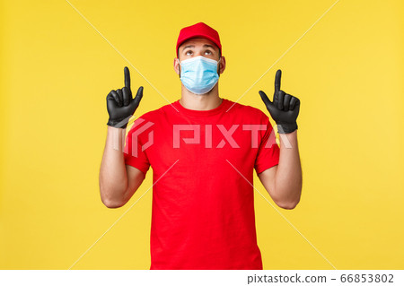 Express delivery during pandemic, covid-19, safe shipping, online shopping concept. Curious young courier in red uniform, medical mask and gloves, looking pointing up, yellow background 66853802