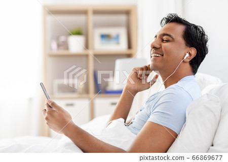 indian man in earphones with phone in bed at home indian man in earphones with phone in bed at home 66869677
