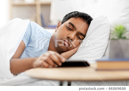 sleepy indian man in bed looking at smartphone sleepy indian man in bed looking at smartphone 66869686