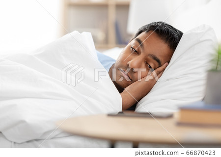 sleepy indian man in bed looking at smartphone sleepy indian man in bed looking at smartphone 66870423