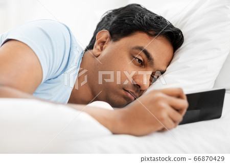 sleepy indian man with smartphone lying in bed 66870429