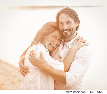 Beautiful couple stand embracing against sea or ocean background. Smiling man and woman in white clothes hugging each other. Tinted image 66880660