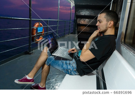 A young man works on a laptop on the deck of a A young man works on a laptop on the deck of a 66891696