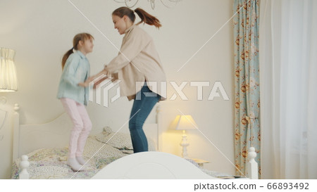 Mother and daughter jumping in bed having fun holding hands enjoying free time indoors 66893492