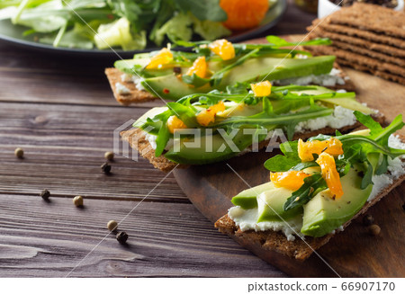 Wholegrain crisp bread with cheese, avocado, green leaves and tangerine on wooden backdrop 66907170