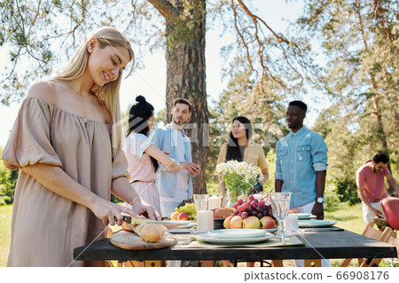 Happy young blond woman cutting fresh baguette on wooden board by table 66908176