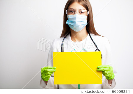 Young female doctor in a mask holds a blank sheet of paper with space for text 66909224
