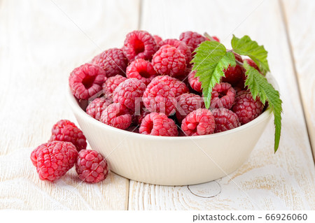 Whole ripe sweet raspberries and green leaf in a beige ceramic bowl on a white wood table. Whole ripe sweet raspberries and green leaf in a beige ceramic bowl on a white wood table. 66926060
