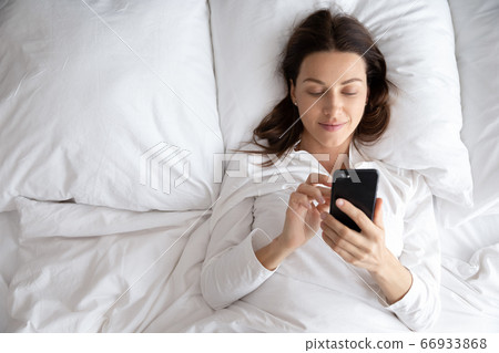 Smiling young woman relax in bed using cellphone Smiling young woman relax in bed using cellphone 66933868