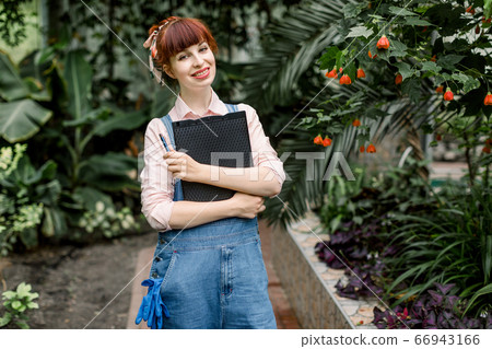 Happy Caucasian woman standing in greenhouse on the background of tropic palm trees and flowers, holding clipboard with notes and looking at camera. Business hothouse start up concept. Happy Caucasian woman standing in greenhouse on the background of tropic palm trees and flowers, holding clipboard with notes and looking at camera. Business hothouse start up concept. 66943166
