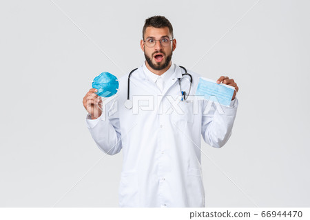 Healthcare workers, medical insurance, pandemic and covid-19 concept. Excited male doctor in glasses and white coat, physician showing medical mask and respirator, gasping amazed 66944470