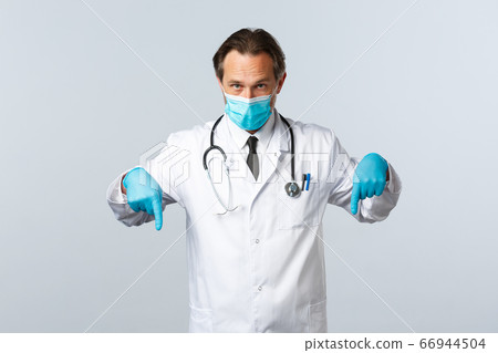 Covid-19, preventing virus, healthcare workers and vaccination concept. Serious doctor in medical mask and gloves, pointing fingers down, showing promo banner, white background 66944504