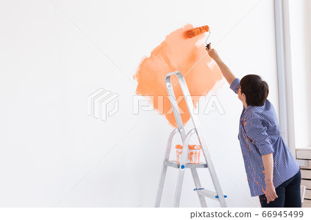 Pretty smiling middle-aged woman painting interior wall of home with paint roller. Redecoration, renovation, apartment repair and refreshment concept. Copy space. Pretty smiling middle-aged woman painting interior wall of home with paint roller. Redecoration, renovation, apartment repair and refreshment concept. Copy space. 66945499