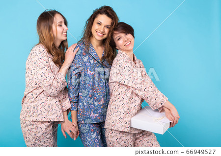 Portrait of three beautiful young girls wearing colorful pyjamas having fun during sleepover isolated over blue background. Pajama party and hen-party concept 66949527
