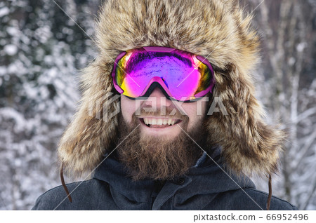 Close-up portrait of a bearded happy snowboarder skier in a ski mask with goggles and a fur big old-school hat on a background of a winter snowy mountains Close-up portrait of a bearded happy snowboarder skier in a ski mask with goggles and a fur big old-school hat on a background of a winter snowy mountains 66952496