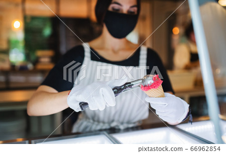 Young woman with face mask working indoors in coffee shop, serving icecream. Young woman with face mask working indoors in coffee shop, serving icecream. 66962854