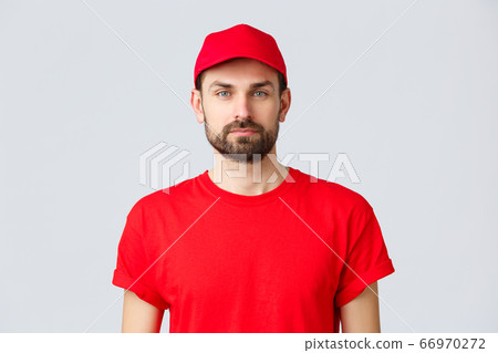Online shopping, delivery during quarantine and takeaway concept. Young handsome bearded courier in red service uniform with cap and t-shirt, employee working transfering orders Online shopping, delivery during quarantine and takeaway concept. Young handsome bearded courier in red service uniform with cap and t-shirt, employee working transfering orders 66970272