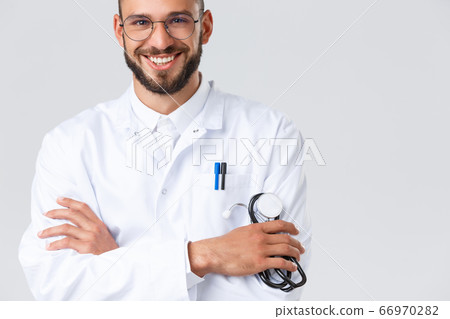 Healthcare workers, coronavirus, covid-19 pandemic and insurance concept. Close-up of handsome smiling male doctor in white coat, glasses, cross arms chest and hold stethoscope, talk to patients 66970282