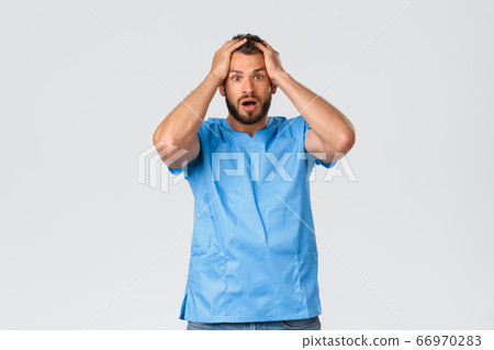 Healthcare workers, medicine, covid-19 and pandemic self-quarantine concept. Shocked and nervous male doctor, nurse in blue scrubs, gasping astonished, hold hands on head startled 66970283