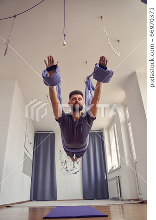 Handsome bearded man practicing aerial yoga in studio Handsome bearded man practicing aerial yoga in studio 66970371