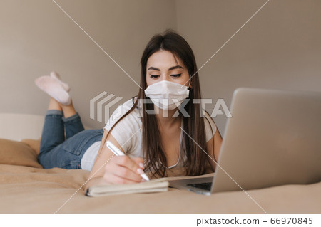 Young woman in mask use laptop on the bed. Female working during quarantine in bedroom. Student study at home. Stay at home 66970845