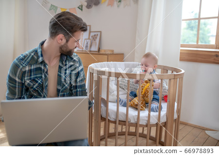Dark-haired father working on a laptop while his little son playing Dark-haired father working on a laptop while his little son playing 66978893