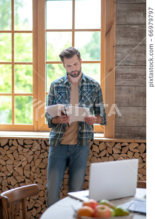 Man in a checkered shirt standing near the window, holding documents and looking concentrated 66979797