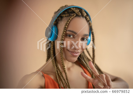 Smiling young African American female in blue headphones, showing peace sign, making selfie Smiling young African American female in blue headphones, showing peace sign, making selfie 66979852