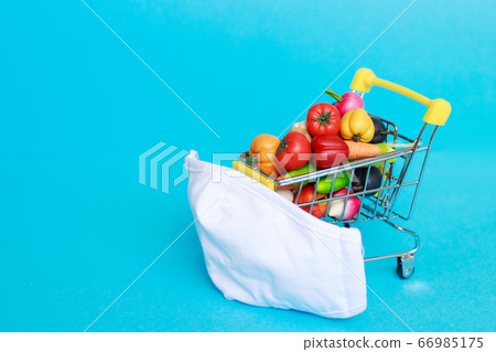 Shopping cart with protective medical mask with fruits and vegetables on a blue background. Safe and online shopping on quarantine concept. Shopping cart with protective medical mask with fruits and vegetables on a blue background. Safe and online shopping on quarantine concept. 66985175