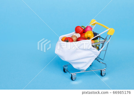 Shopping cart with protective medical mask with fruits and vegetables on a blue background. Safe and online shopping on quarantine concept. Shopping cart with protective medical mask with fruits and vegetables on a blue background. Safe and online shopping on quarantine concept. 66985176