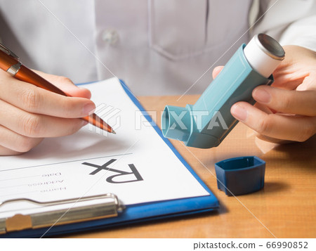 Close up of doctor hand holding blue asthma inhaler and writing medical prescription on rx form for treatment asthma or COPD diseases on physician's desk at hospital. Healthcare concept. 66990852