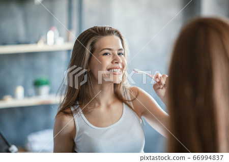 Daily morning routine. Millennial girl with toothbrush cleaning her teeth near mirror indoors Daily morning routine. Millennial girl with toothbrush cleaning her teeth near mirror indoors 66994837
