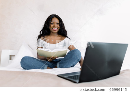 Young african american girl in white t-shirt and jeans, sitting on bed with laptop pc and notepad in bedroom, studying or working at home, spending time at home. Online work concept 66996145