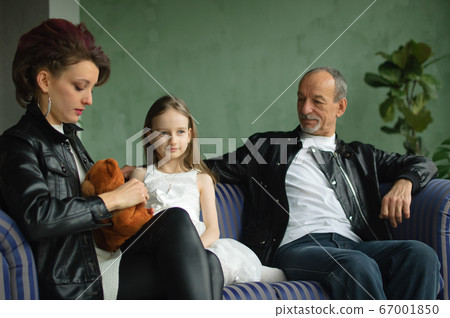 Family portrait of adult daughter, little granddaughter and senior grandfather in loft room with houseplants. Man and woman are wearing black leather jackets in punk style, generation concept 67001850