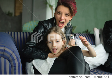 Crazy mother in black leather jaket and pants hugging her little daughter in white dress, they are looking at the camera with opened mouths and grimacing in loft room with houseplants. Crazy mother in black leather jaket and pants hugging her little daughter in white dress, they are looking at the camera with opened mouths and grimacing in loft room with houseplants. 67001851