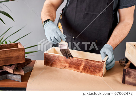 Man carpenter varnishing wooden crate for flowers with brush in her small business woodwork workshop. In your work, do you use stains or wood preservatives to show the wood pattern. 67002472