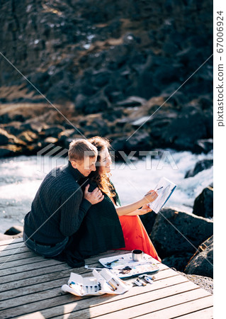 A guy hugs a girl artist who paints a picture in the album with watercolors. Near Ehsaraurfoss waterfall, Ehsarau River, Thingvedlir National Park in Iceland. 67006924