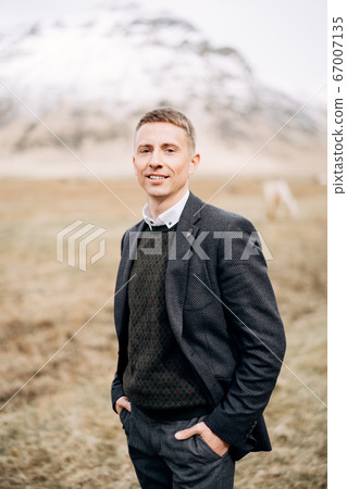 Destination Iceland wedding. Close-up man portrait. A guy in a sweater in a rhombus, a black jacket, stands in field of dry grass, holding his hands in his pockets, against backdrop of snowy mountain 67007135