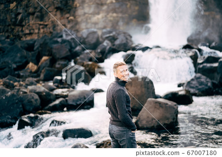 A guy in a black knitted sweater stands on rocks near Ehsaraurfoss Falls, Ehsarau River, National Park, Tingvedlir, Syudurland Region, Iceland. A guy in a black knitted sweater stands on rocks near Ehsaraurfoss Falls, Ehsarau River, National Park, Tingvedlir, Syudurland Region, Iceland. 67007180